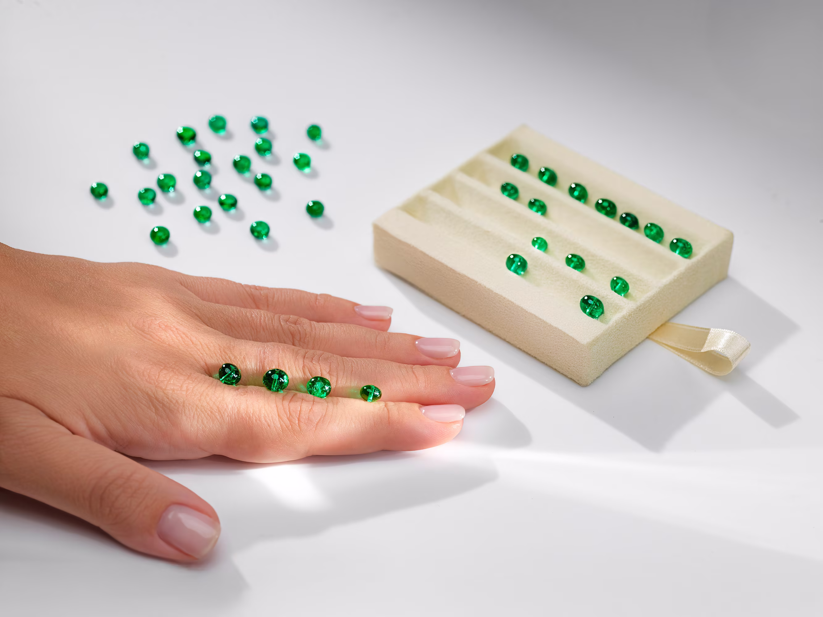 Hand on table with green emeralds resting on fingers; others scattered nearby and some lined up on a light velvet backing.