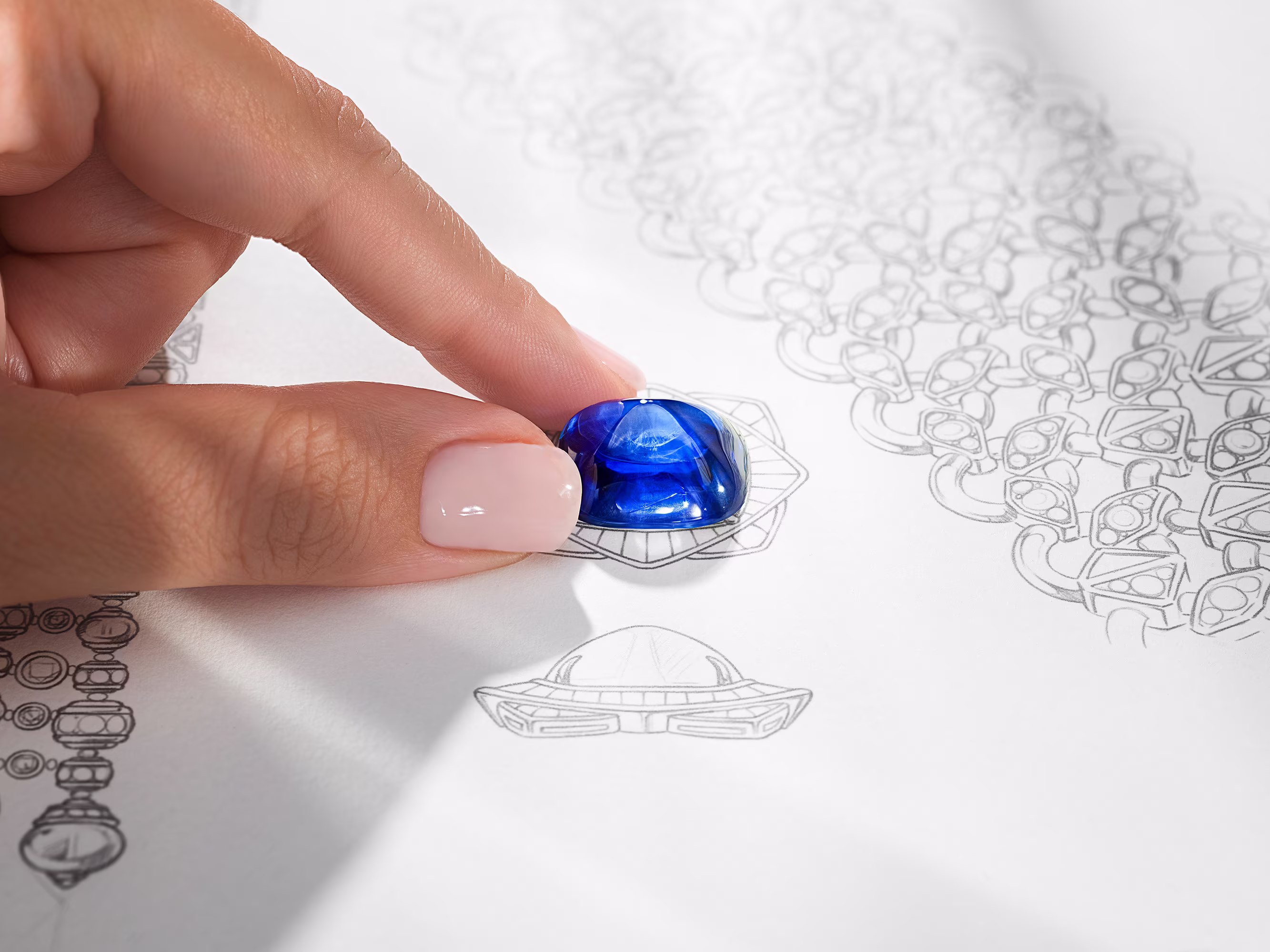 A hand with manicured nails holds a blue sapphire on a technical drawing of jewelry with geometric motifs.