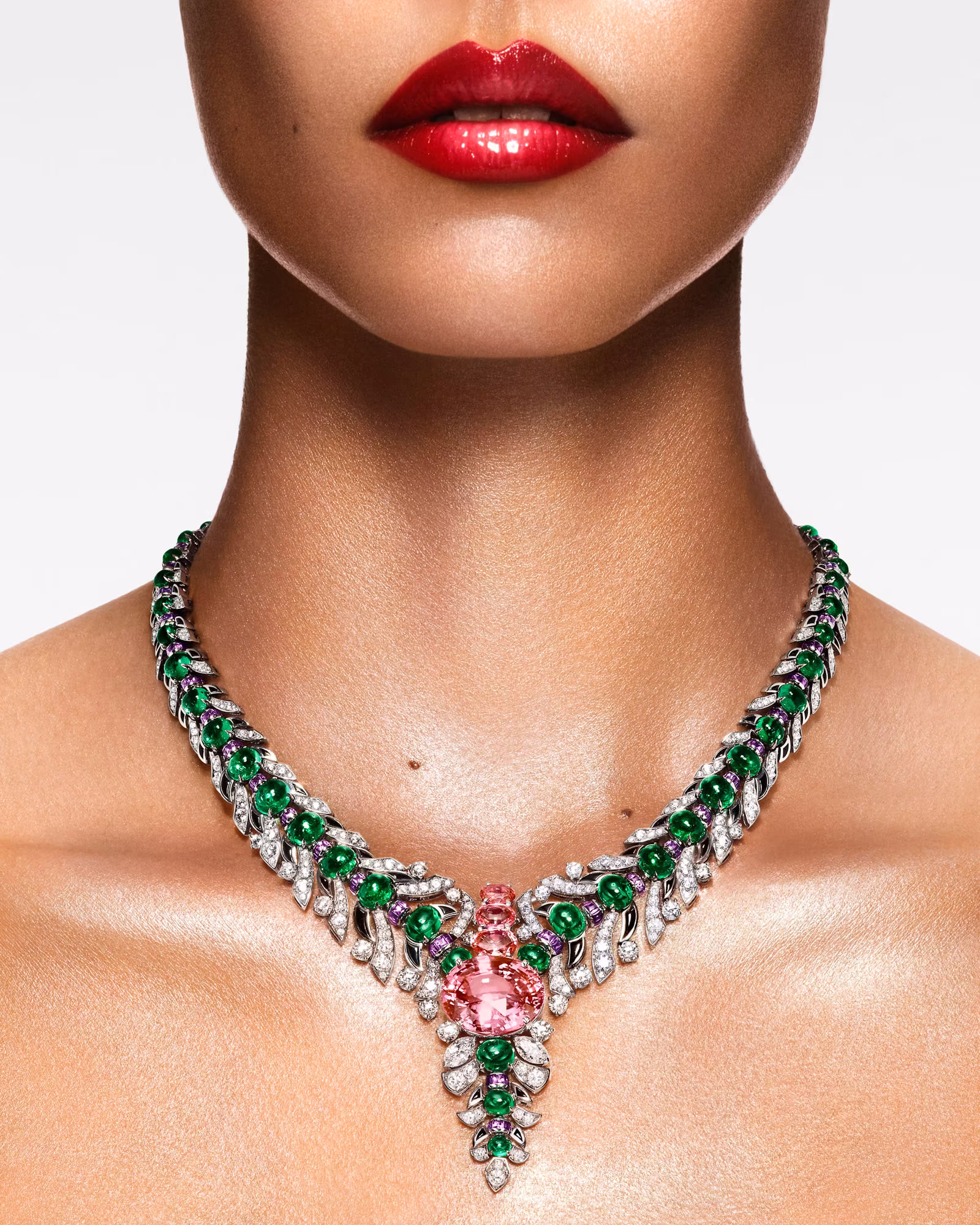 Close-up of neck with white gold necklace, diamonds, and green emeralds with a large pink stone in the center. Red lips against a light background.
