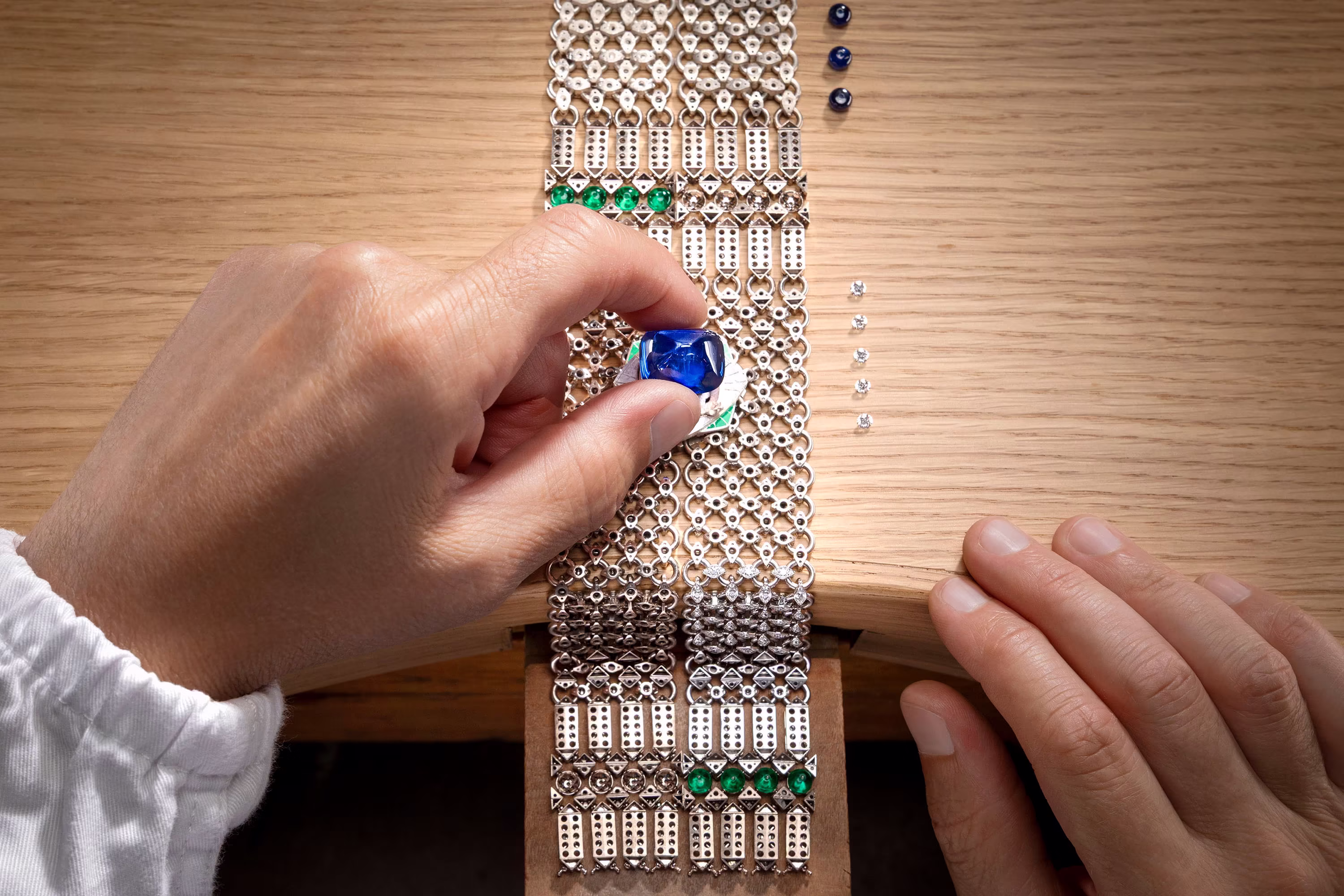 Hands place a large blue sapphire onto a diamond and emerald necklace during jewelry setting on a wooden workbench.