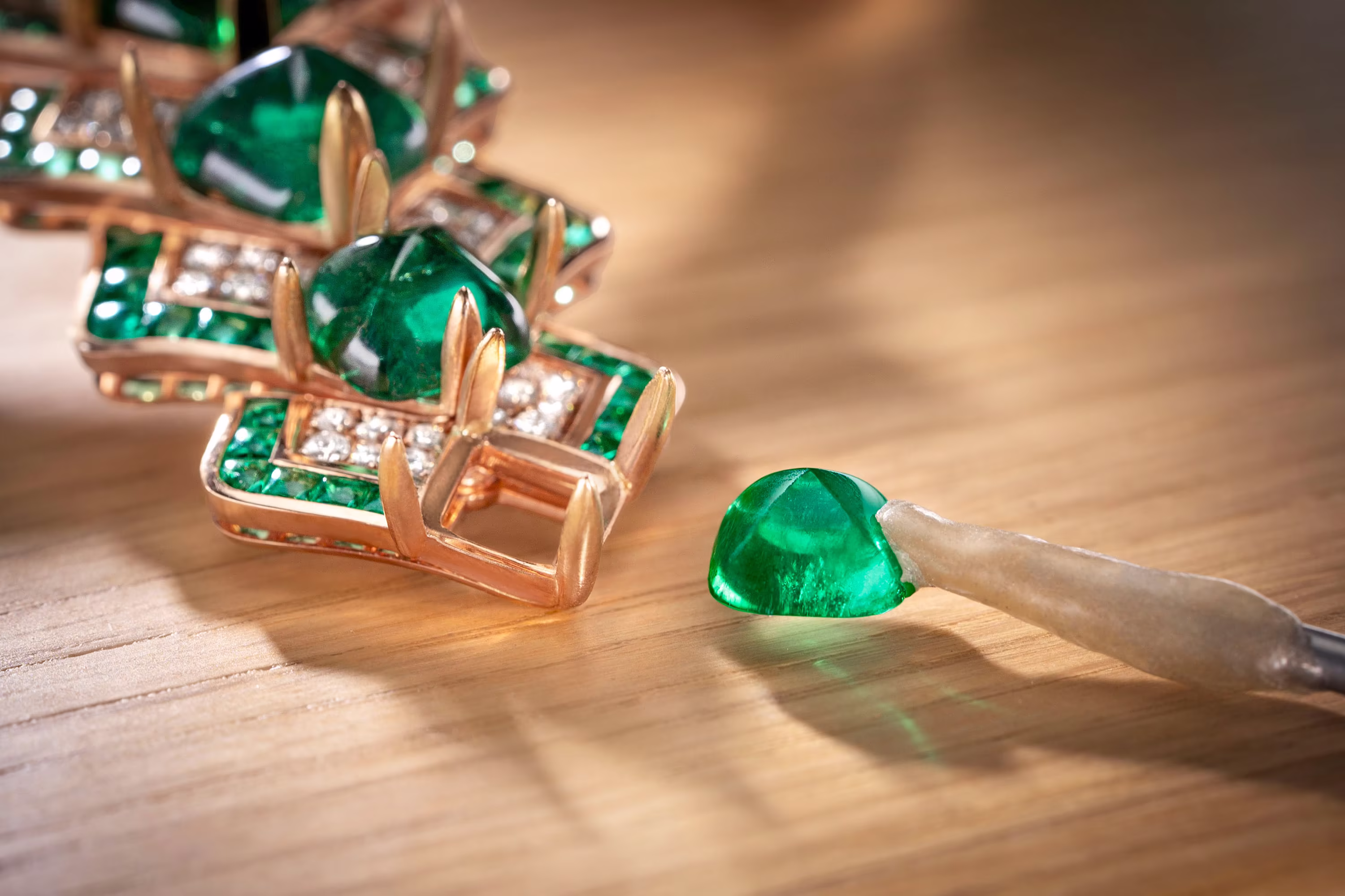 Loose green gemstone beside a gold jewelry setting with diamonds, while a tool prepares the stone for mounting on a wooden workbench.