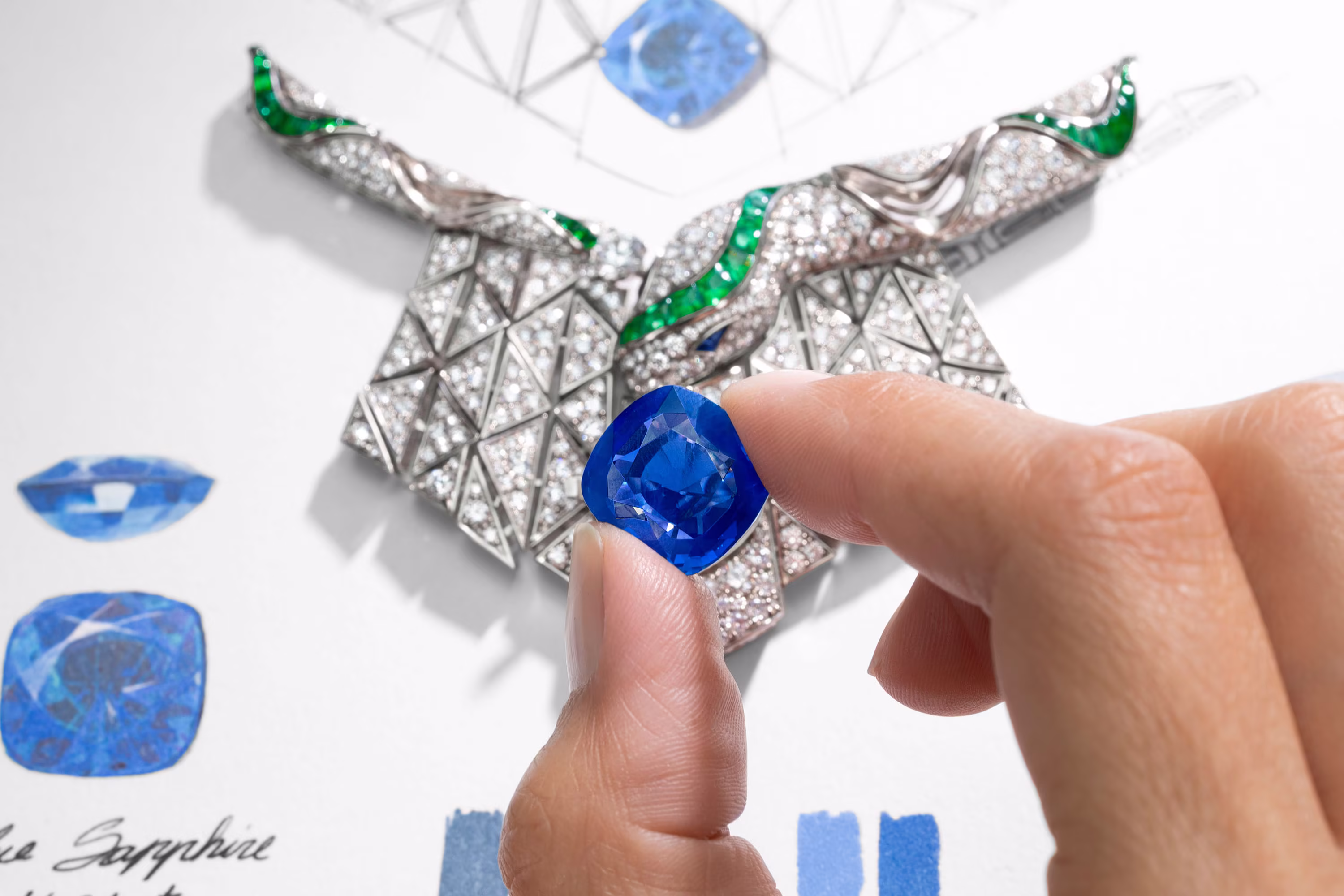 Mano holds a blue gemstone above the design of a necklace with diamonds and green details, evaluating the setting.