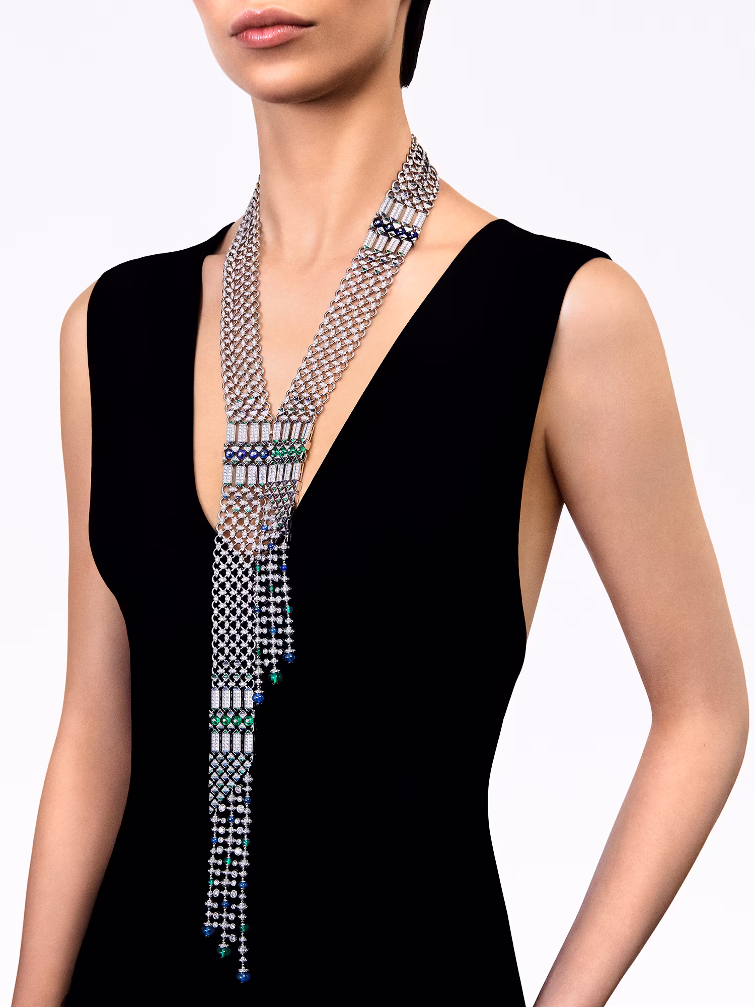 Model in a black dress wears a long necklace with diamonds, emeralds, and blue stones that falls down her chest.