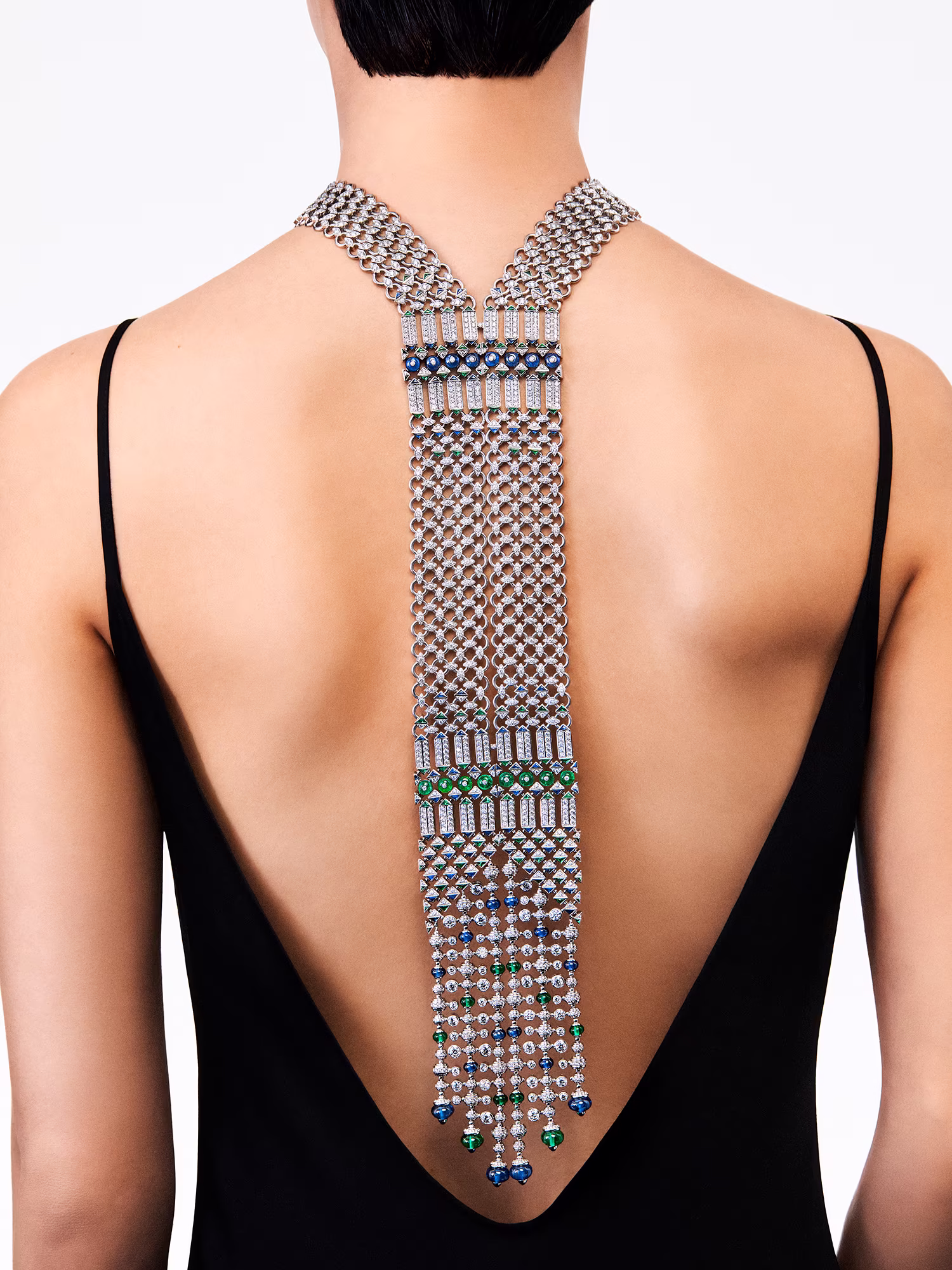 Rear view: model wearing a low-cut black dress with a long necklace featuring diamonds, emeralds, and blue stones on her back.