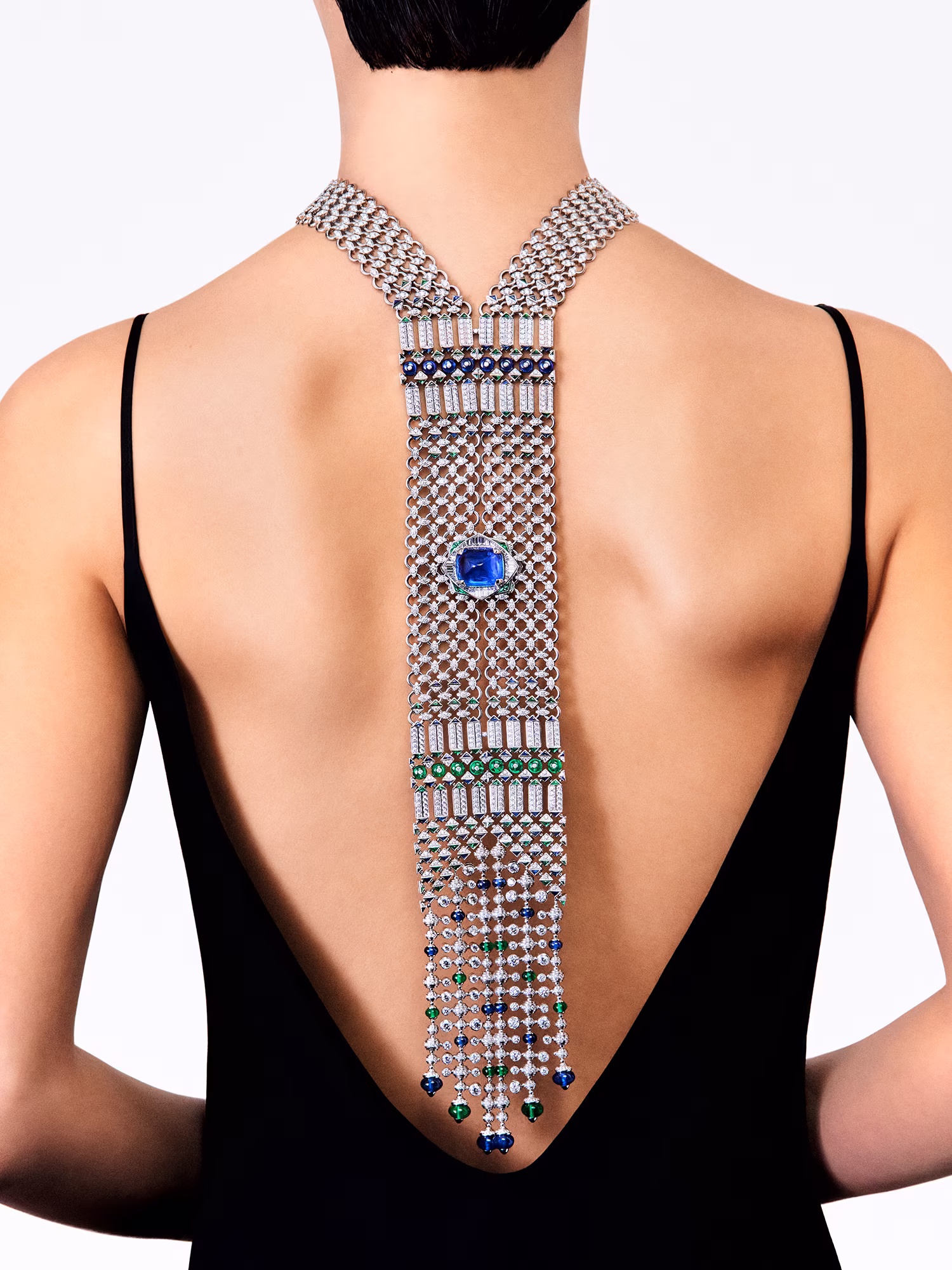 Rear view: model wearing a low-cut black dress with a long necklace featuring diamonds and blue and green stones on her back.
