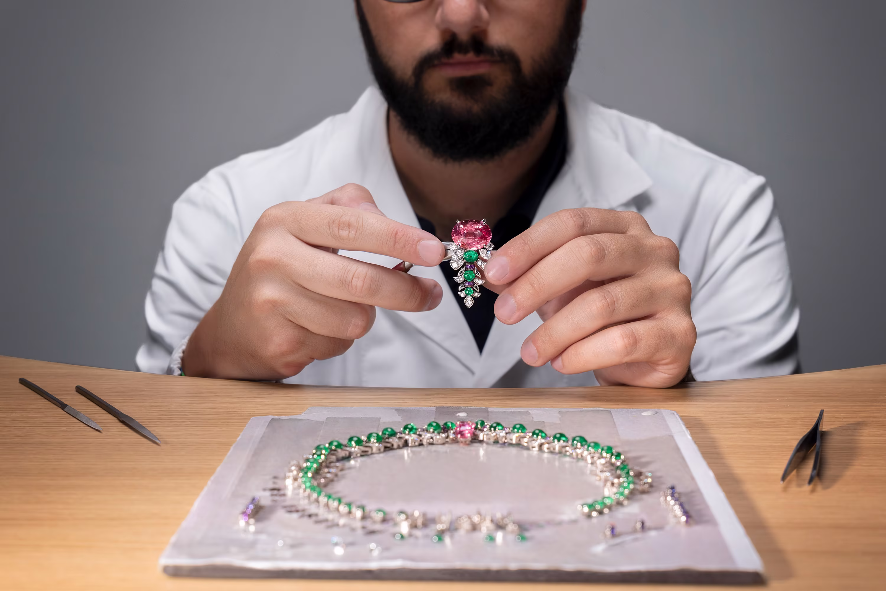 Craftsman at workbench holds pendant with pink stone and emeralds above necklace in progress. Tools on table.
