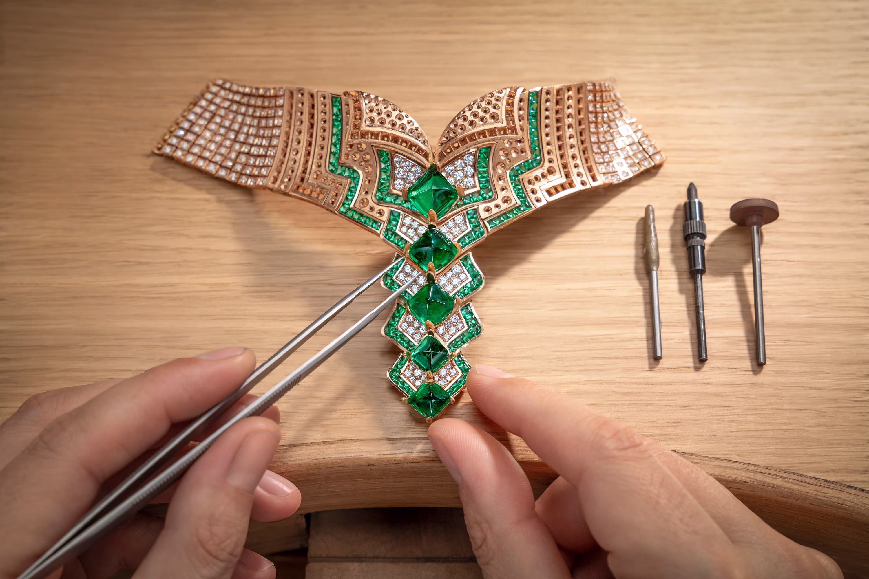 Jeweler adjusting emerald stones in a gold necklace with tweezers on a wooden bench, polishing tools neatly placed beside.