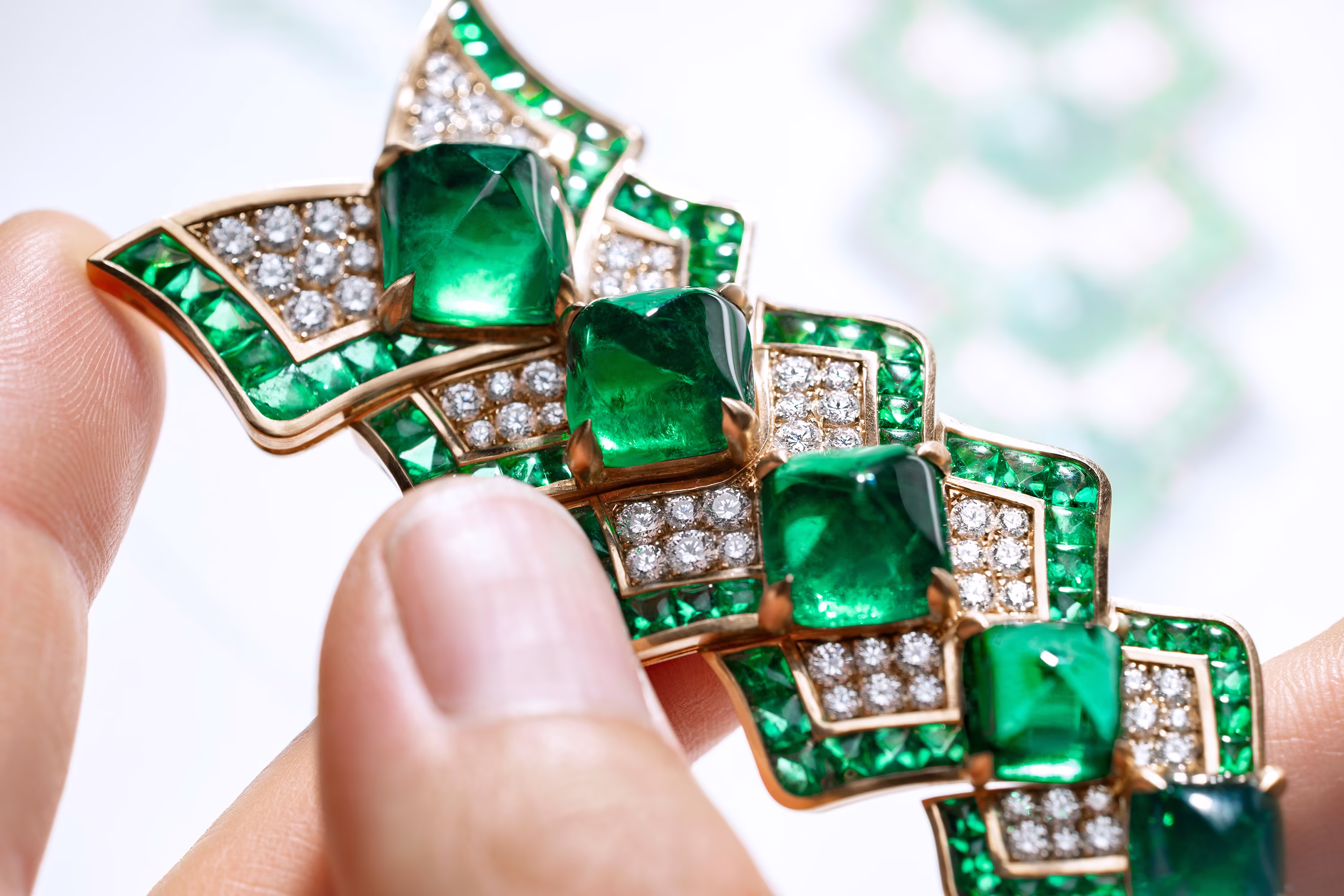 Close-up of emerald and diamond necklace held between fingers, showing vivid green stones set in gold with sparkling details.