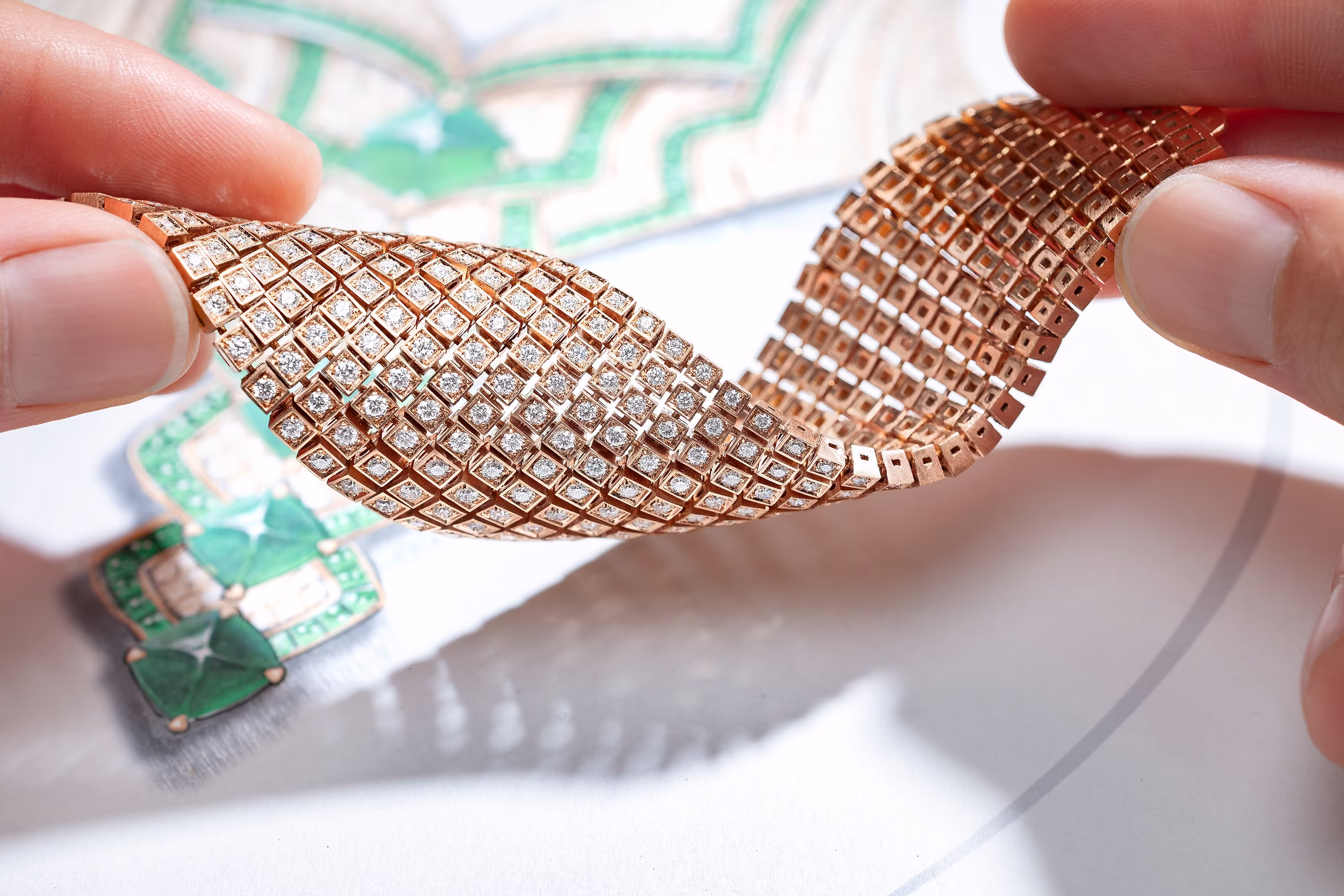Hands holding a flexible diamond bracelet in rose gold above a jewelry design sketch with green gemstones in the background.