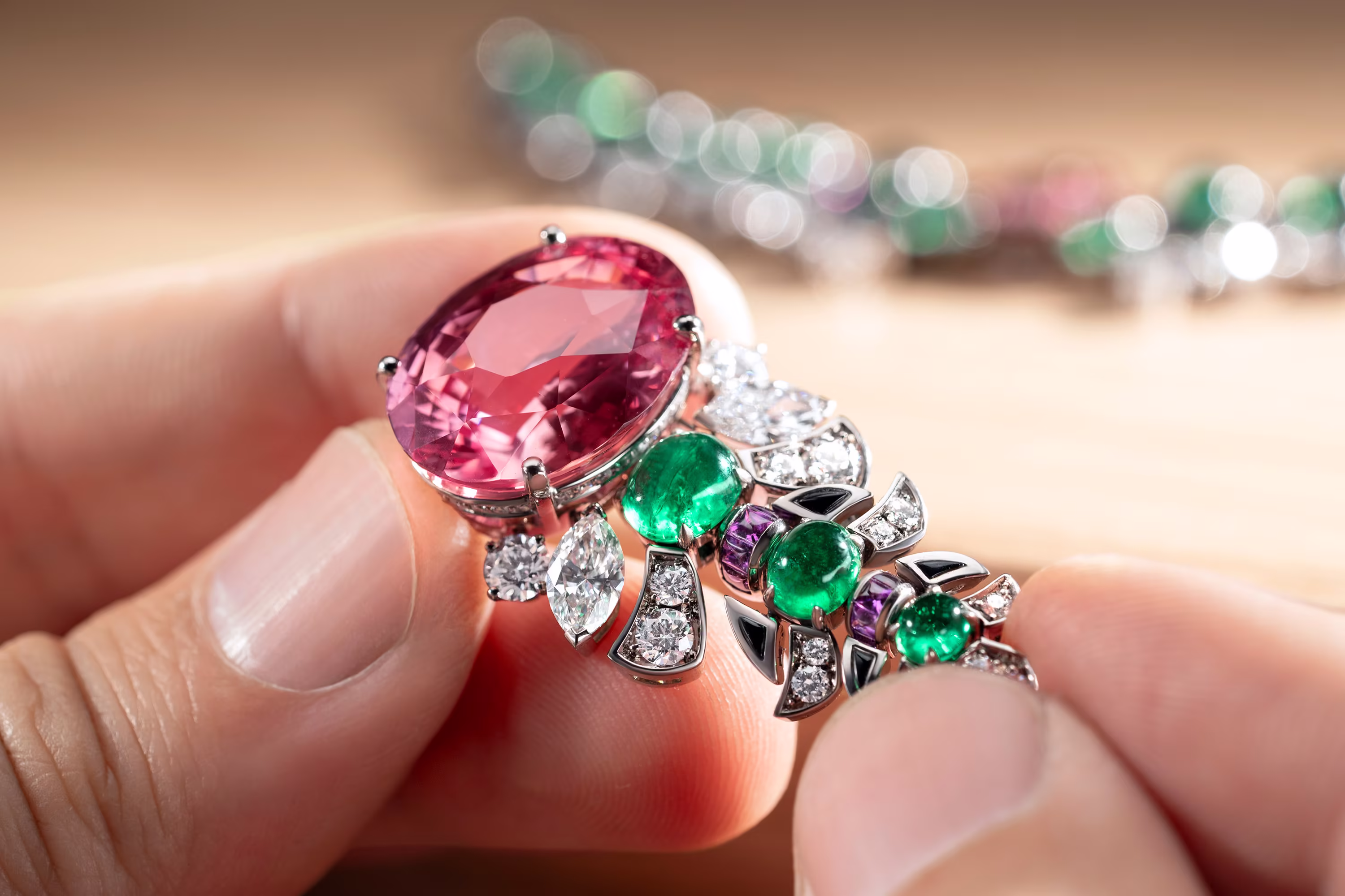 Hands hold a pendant with a large pink stone, green emeralds, and diamonds; in the background, a blurred necklace lies on the goldsmith's bench.