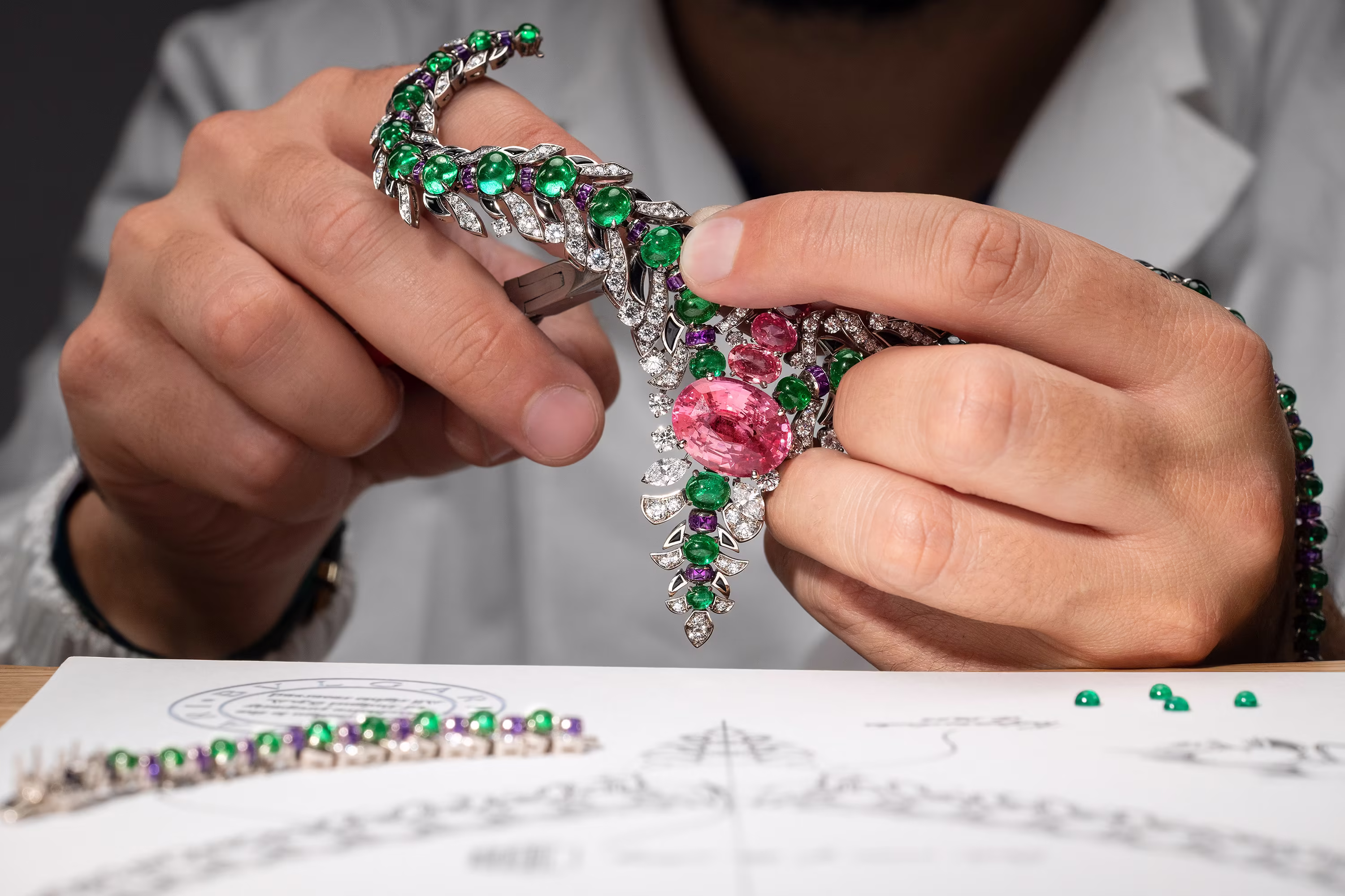 Goldsmith's hands work on a necklace with emeralds, diamonds, and a large pink stone above the technical drawing of the jewelry.