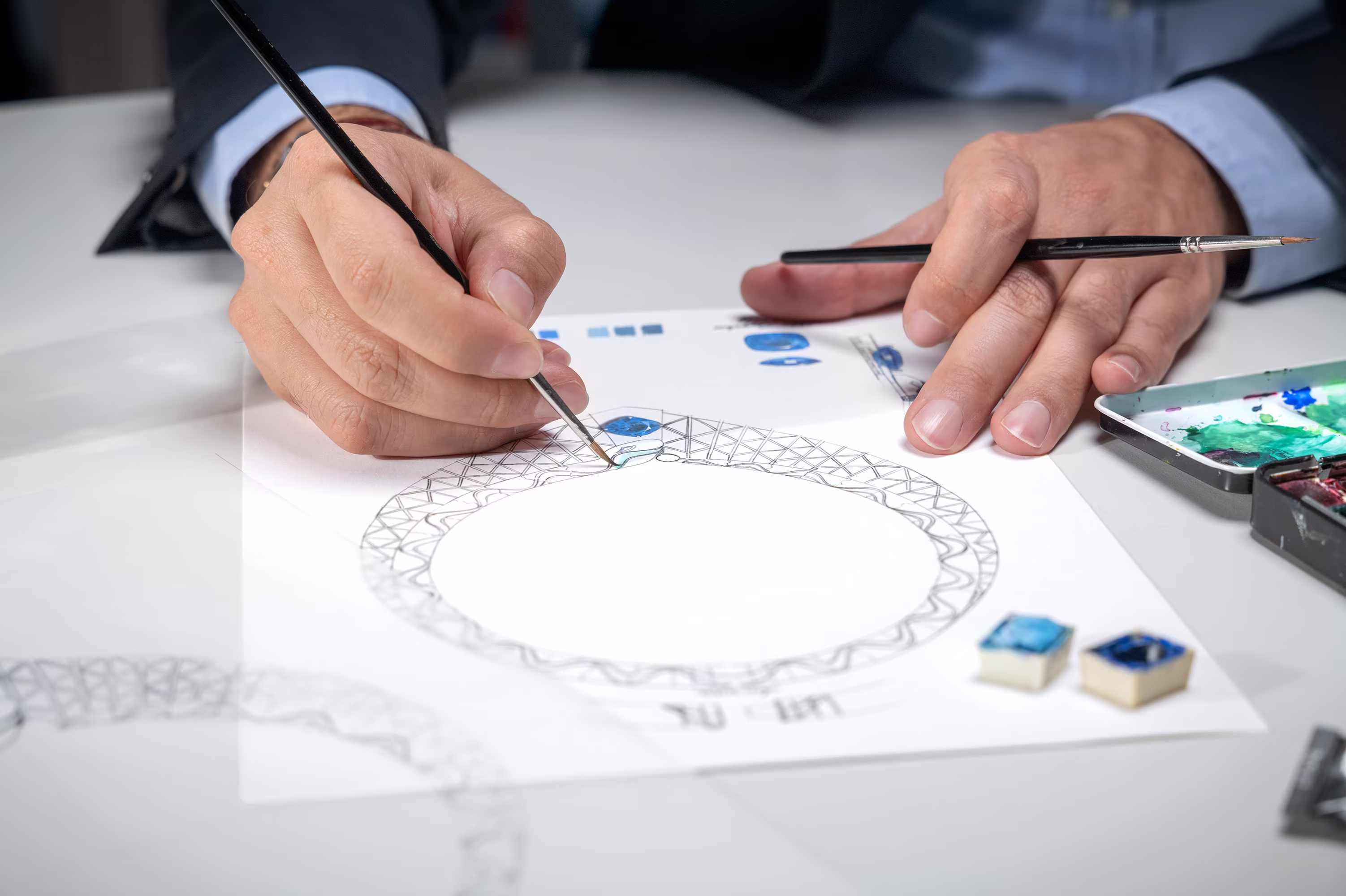 Hands paint the design of a necklace with a blue stone in the center; next to it, colors and gem samples on the table.