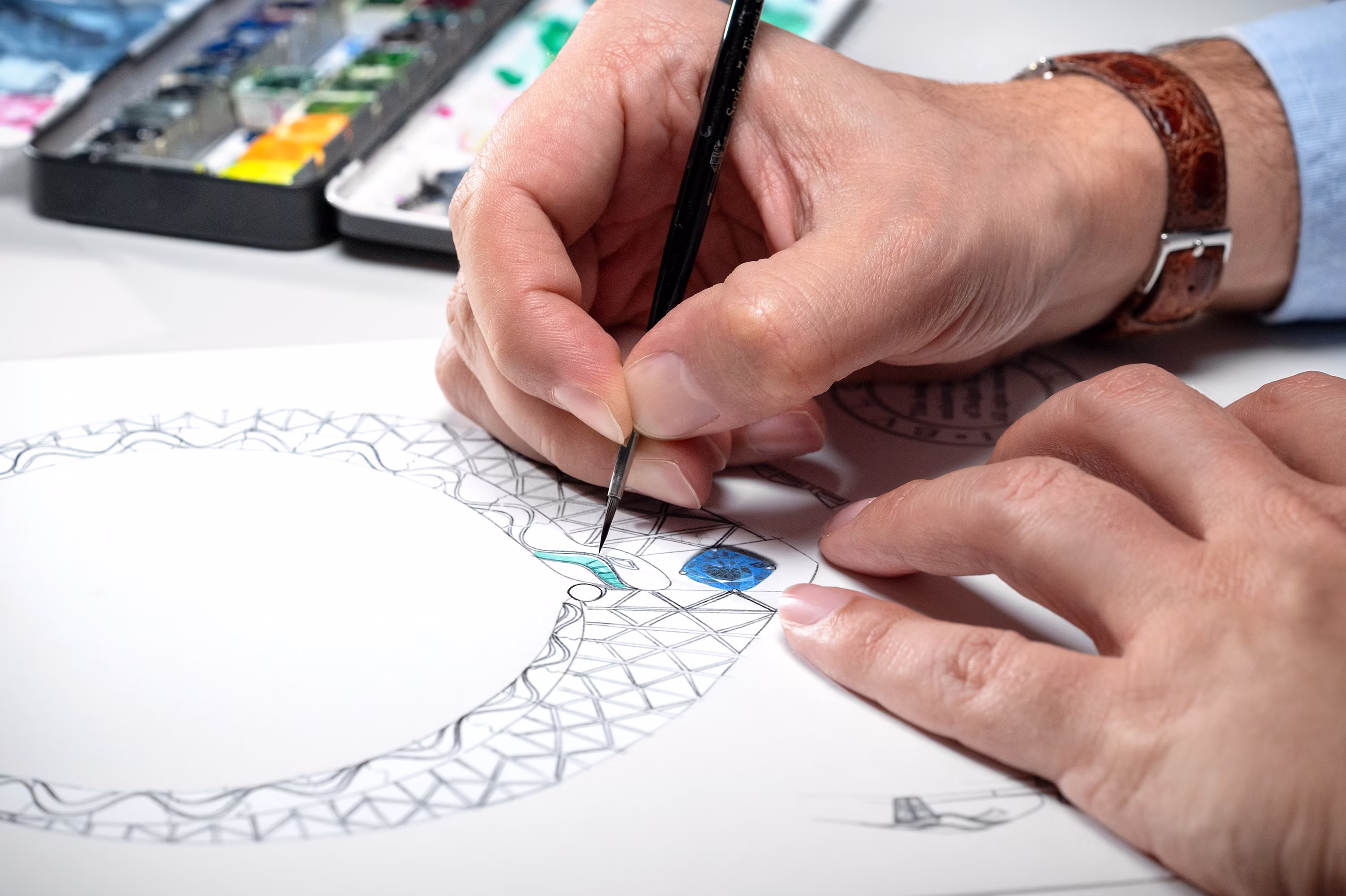 Hands paint the design of a necklace with a blue stone in the center; on the table are a palette and paints.