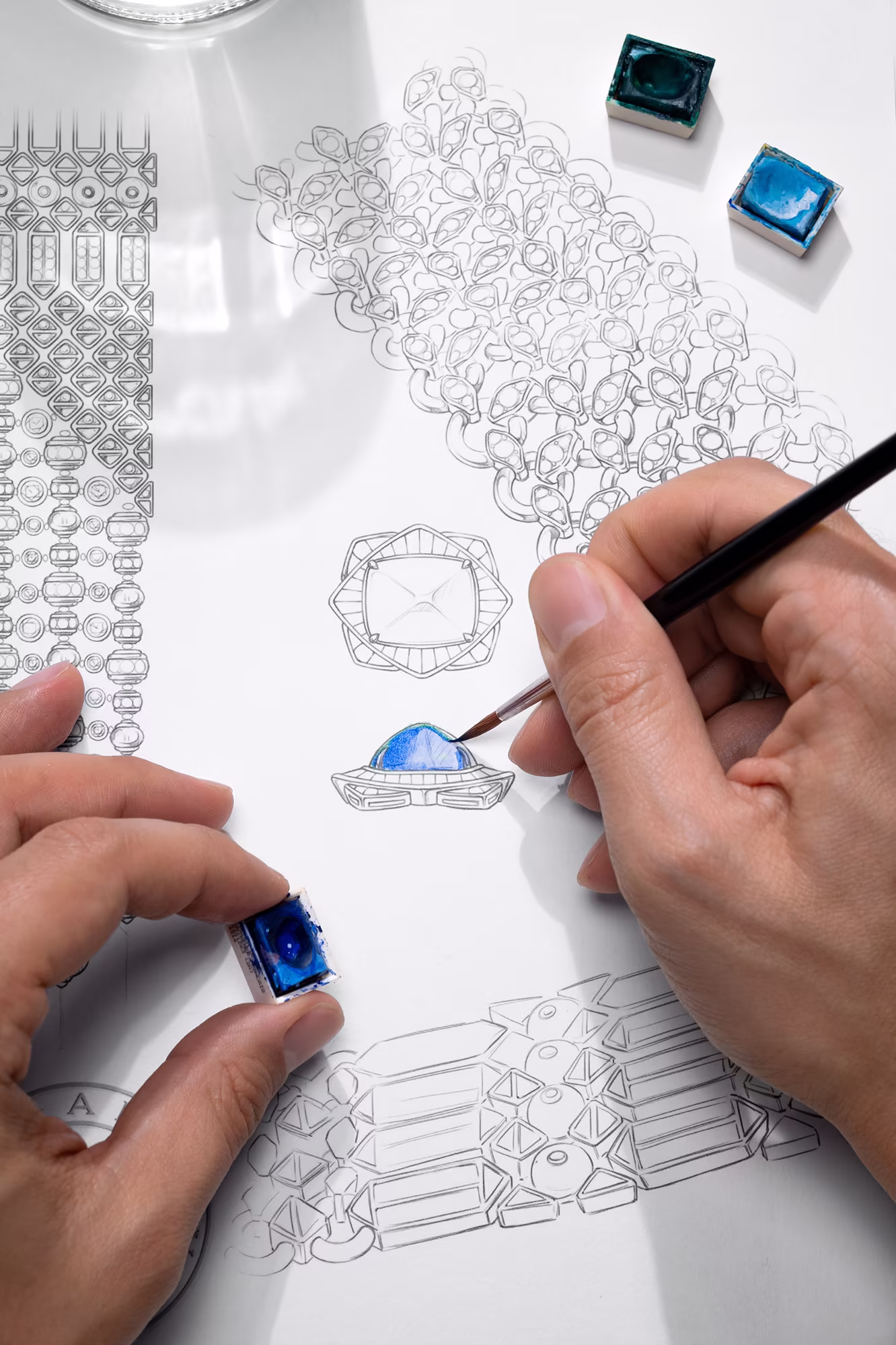 Hands paint a blue gemstone detail on a jewelry design sketch while holding a sapphire color sample nearby.