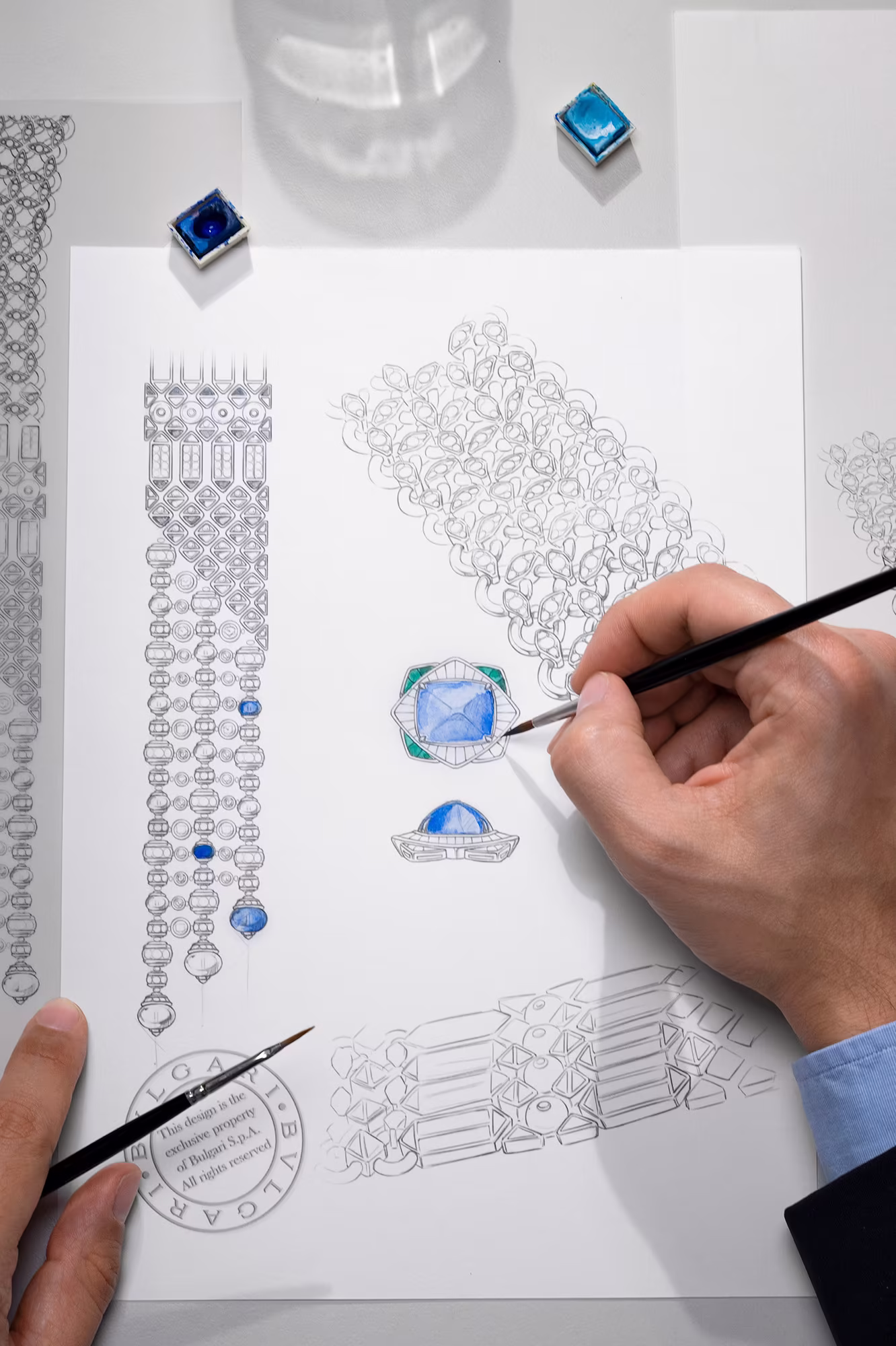 Hand painting a blue gemstone on a detailed jewelry design sketch, with tools and color samples on the worktable.