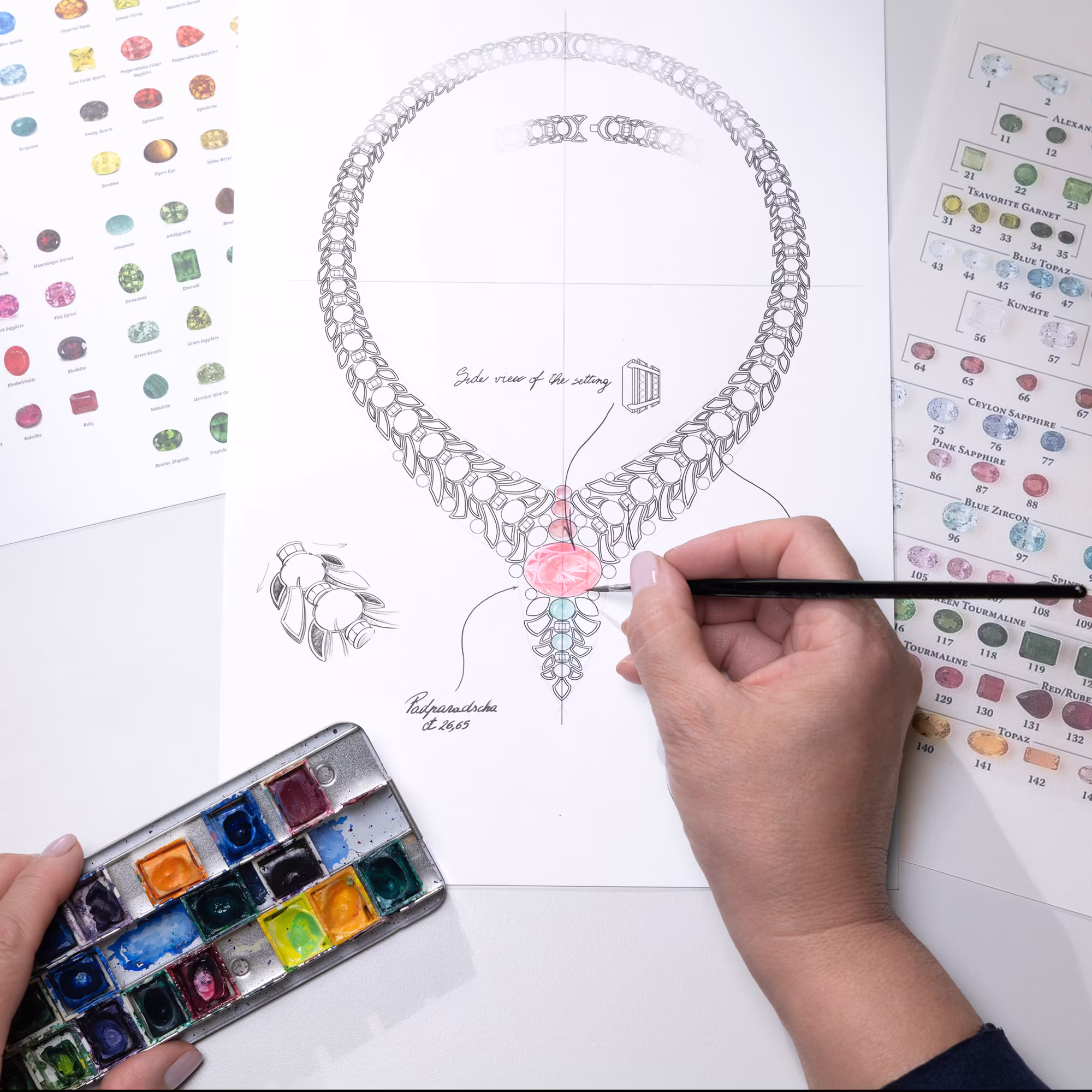 Mano paints the design of a necklace with a large pink stone in the center; next to him are a color palette and boards with gems.